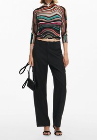 Femme portant un pantalon noir à jambes larges, des talons ouverts et un haut à manches longues rayé multicolore, tenant un petit sac à main noir.