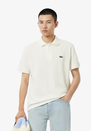 Witte katoenen polo shirt met een puntige kraag en korte mouwen, met een klein blauw Lacoste-logo op de borst. Lichtblauwe spijkerbroek.