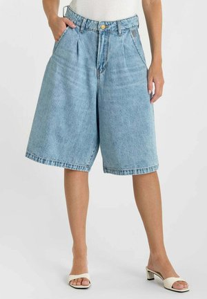 Donna che indossa pantaloncini di denim azzurro chiaro, ampi, lunghezza al ginocchio, con pieghe frontali e sandali bianchi con tacco, su sfondo semplice.
