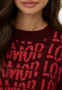 Pull bordeaux avec motif texte "L O V E" en rouge, col rond, texture tricotée, gros plan sur le haut du corps et l'encolure sans détails faciaux.