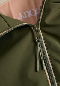 Primo piano di una cerniera di una giacca verde oliva con due tiranti e il marchio "JJXX" sul tessuto e sul cursore della cerniera.