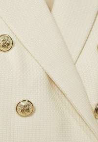 Kaksirivinen bleiseri off-white värissä, jossa on teksturoitu kangas ja kultaiset leijonapäät painikkeet. Sen terävät kaulukset ja räätälöity istuvuus luovat eleganssia.