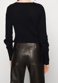 Personne portant un pull en maille côtelée noire et un pantalon en cuir noir taille haute, vue de dos sur un fond uni.