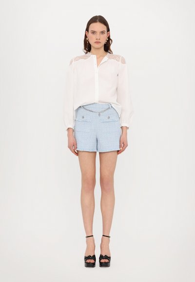 Camisa blanca de botones con detalles de encaje en los hombros, combinada con pantalones cortos de tweed azul claro con bolsillos delanteros y un cinturón de cadena. Tacones negros.