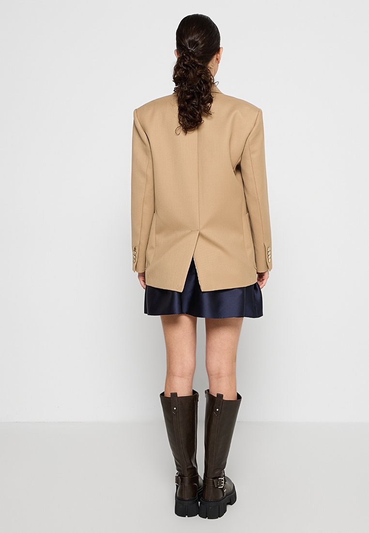 Beige oversized blazer met een split aan de achterkant, gedragen over een marineblauwe satijnen rok, gecombineerd met kniehoge bruine leren laarzen met gespen.