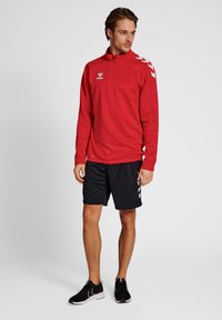 Giacca sportiva rossa con zip e colletto alto, pantaloni corti neri da atleta e sneaker neri. La giacca presenta strisce bianche sulle maniche.