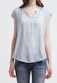 Světlomodrý top s krátkým rukávem a krajkovými detaily na ramenech, V-výstřihem a volným střihem. Tkanina vypadá měkce a má mírný lesk.