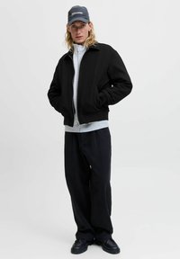 Zwarte bomberjack met een grijze rits-hoodie eronder, gecombineerd met losse zwarte broek en zwarte schoenen, verfraaid met een pet.