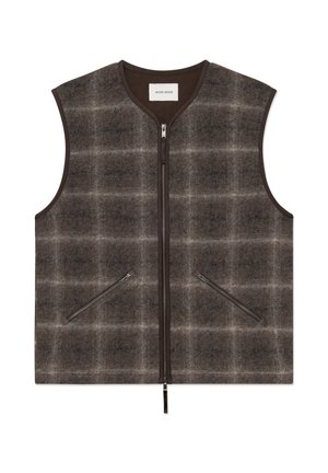 Homme portant une casquette noire, un gilet marron à carreaux sur une chemise rayée foncée, un pantalon noir et des chaussures noires, debout les mains dans les poches du gilet sur fond blanc.