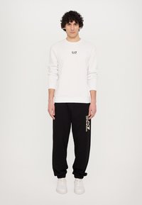 Hvid langærmet sweatshirt med logo, parret med sorte sweatpants med grafisk tryk i hvid, afsluttet med hvide sneakers.