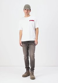 Bílé tričko s krátkým rukávem a červeným logem, v kombinaci se slim-fit šedými džíny a béžovými šněrovacími botami. Čepice je olivově zelená s vyšívaným textem.