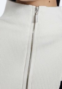 Gros plan sur une fermeture éclair argentée sur un vêtement blanc côtelé à col montant, partiellement fermée près du cou de la personne.