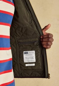 Hånd, der holder en mørk quiltet jakke åben og afslører en lynlåslomme og en stofetiket med tekst og et britisk flag.