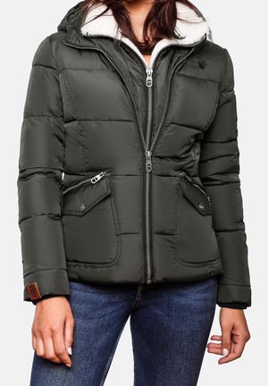 Kobieta w ciemnozielonej pikowanej kurtce z kapturem wyściełanym białym polarem oraz niebieskich dżinsach, stojąca z rękami swobodnie opuszczonymi wzdłuż ciała.