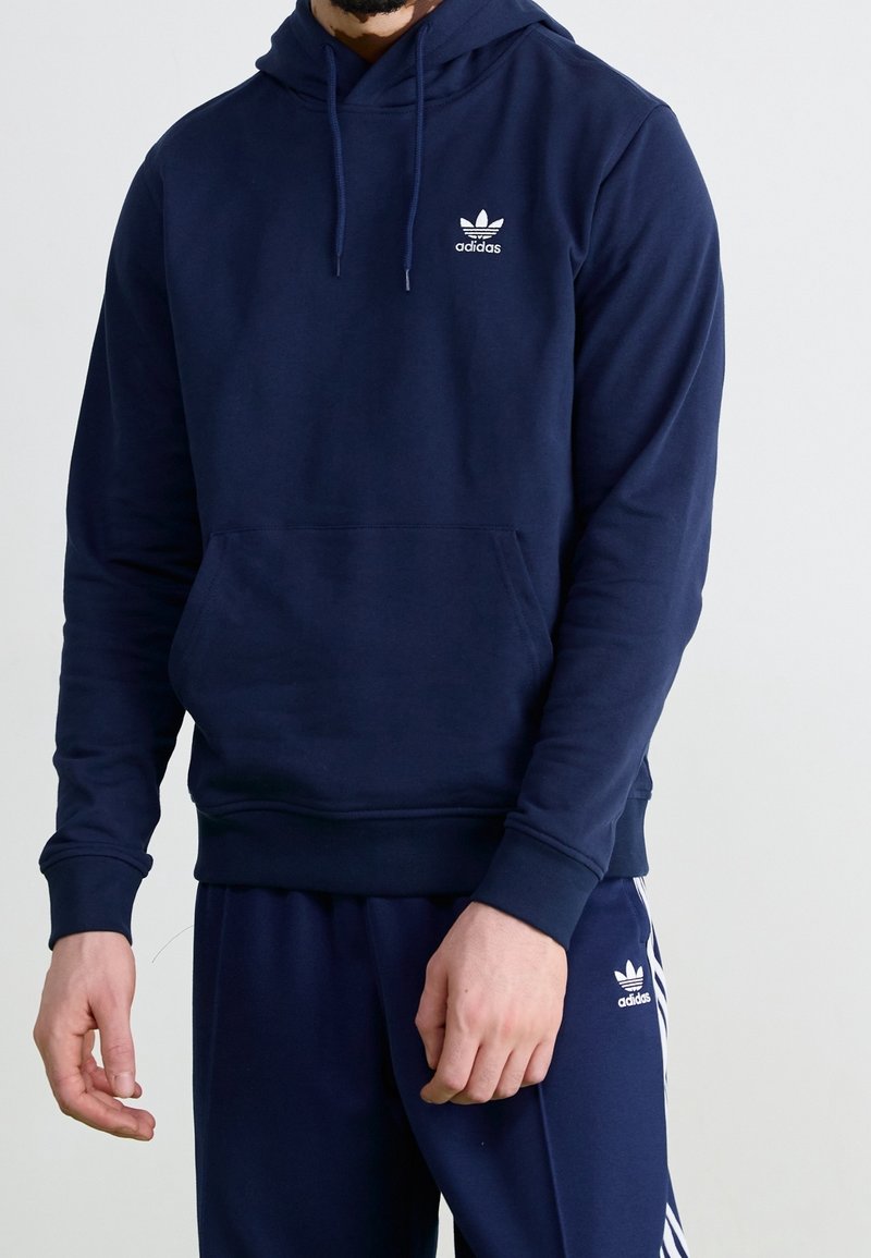 Mörkblå hoodie med känguruficka, ribbade ärmslut och nederkant, samt vitt Adidas-logotyp på vänster bröst. Tillverkad av mjukt, texturerat tyg.