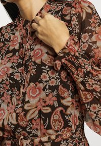 Kukallinen mekko, jossa on tumma ruskea tausta, vaaleanpunaisia ja kermaisia kukkia, pitkät hihat, joissa on kerrokselliset hihansuut sekä nauhakiinnitys kauluksessa.