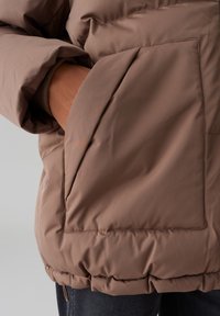 Doudoune marron avec une grande poche avant, des poignets côtelés et un ourlet élastiqué froncé, dotée d'un extérieur lisse et résistant à l'eau.