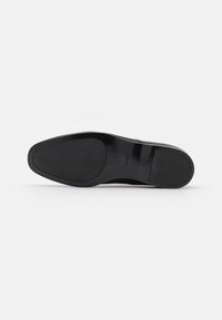 Semelle de chaussure habillée noire avec des grips en caoutchouc texturés au talon et à l’avant, affichant le nom de la marque Aldo près de la voûte plantaire.