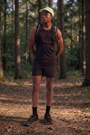 Een atletisch persoon staat op een bospaadje met zwarte short, zwart tanktop, lichtgroene pet, zwarte sokken, hardloopschoenen en een hydratatievest.