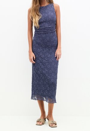 Femme portant une robe midi bleu marine texturée sans manches avec des sandales beiges à bout ouvert, debout sur un fond blanc uni.
