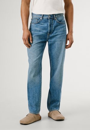 Mann trägt hellblaue Jeans mit geradem Schnitt, weißes T-Shirt und beige Slipper, steht vor einem schlichten Hintergrund.
