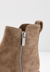 Beżowy zamszowy botek za kostkę z bocznym zamkiem błyskawicznym, charakteryzujący się gładką fakturą, zaokrąglonym szczytem i małą pętelką ułatwiającą zakładanie.