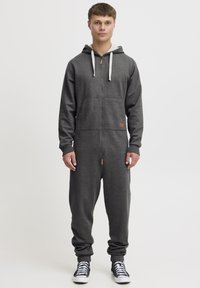 Tmavo šedý overal na zips s kapucňou, ktorý má dve predné vrecká, rebrované manžety a malý akcent loga. Materiál vyzerá mäkký a textúrovaný.