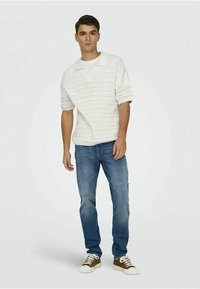 Maglione bianco a maglia con motivo testurizzato, indossato sopra una t-shirt bianca. Abbinato a jeans blu in denim e sneaker marroni. Modello in piedi su uno sfondo chiaro.
