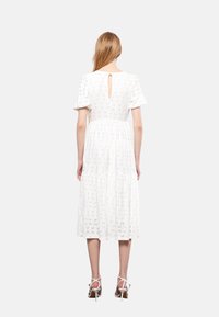 Kobieta z długimi blond włosami, ubrana w białą sukienkę midi z bufiastymi rękawami i dekoltem w kształcie łezki, w białych sandałach na wysokim obcasie, stojąca.