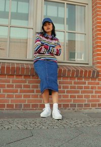 Striped long-sleeve shirt, blue denim skirt, white sneakers, wearing a blue cap; standing against a brick wall with a window behind.
