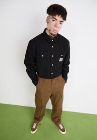 Schwarze Hemdjacke mit zwei vorderen Taschen und Logopatch, kombiniert mit braunen Cargo-Hosen und braunen Sneakern mit weißen Schnürsenkeln.