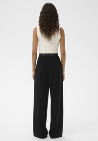 Vrouw met lang krullend haar, gekleed in een mouwloos wit topje dat is ingestopt in een zwarte, hooggetailleerde broek met wijde pijpen en een zwarte riem, achteraanzicht.