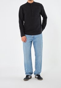 Camicia nera a manica lunga con colletto henley con bottoni e taschino sul petto, abbinata a jeans blu chiaro a vestibilità morbida e sneakers nere con strisce bianche.
