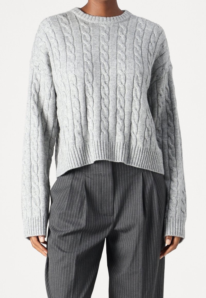 Pull en maille torsadée gris clair avec un col rond et des poignets côtelés, longueur courte, porté sur un pantalon gris à fines rayures.