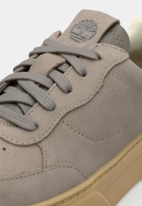 Sneaker din suede bej cu șireturi gri, cu o limbă texturată și o talpă din cauciuc maro; logo-ul este embosat pe limbă.