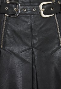 Černá kožená sukně s širokým páskem a kovovou sponou, odhalenými zipy a úhlovými sklady na textuře látky.