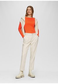 Witte wollen broek met een rechte pijp, gecombineerd met een oranje langemouwen top en een crèmekleurige vest over de schouders gedrapeerd. Beige enkellaarzen.