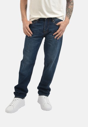 Uomo con jeans blu scuro, sneakers bianche e t-shirt bianca con tatuaggi su entrambe le braccia, in piedi con le mani in tasca.