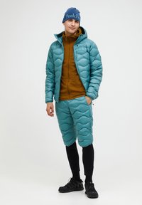 En teal farvet pufferjakke med bølge mønster, en brun hoodie med lynlås, teal shorts og sorte leggings. Fuldfører looket med sorte sneakers og en blå hue.