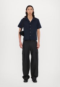 Kurzärmliges navy Hemd mit Knöpfen und einer Brusttasche; kombiniert mit schwarzen Cargo-Hosen mit mehreren Taschen. Das Modell steht aufrecht mit einer Tasche.