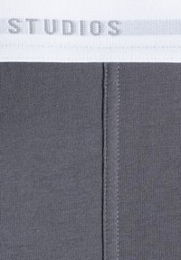 Gray waistband featuring embroidered "STUDIOS" text, stretchy fabric with a smooth texture, and flat seams. Simple design with no additional patterns.