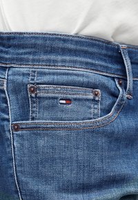 Τσέπη μπλε τζιν με μικρό κόκκινο, λευκό και μπλε ορθογώνιο λογότυπο ραμμένο στην τσέπη για κέρματα, φορεμένη με λευκό πουκάμισο που είναι μπουκωμένο μέσα.