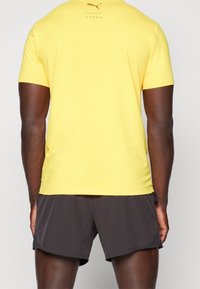 Camiseta atlética amarilla de manga corta y cuello redondo, con un logo marrón y texto "HYROX" en la parte trasera, combinada con pantalones cortos oscuros.