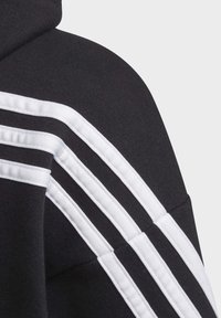 Felpa nera con cappuccio, caratterizzata da tre strisce bianche lungo la spalla. Realizzata in un tessuto testurizzato con una finitura morbida. Cappuccio regolabile.