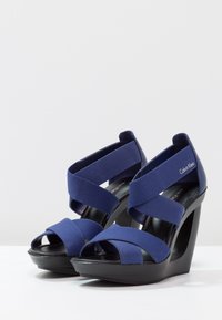 Sandales compensées en tissu bleu avec un design à bout ouvert, présentant des lanières croisées et un talon plateforme noir élégant. Nom de la marque visible sur le côté.