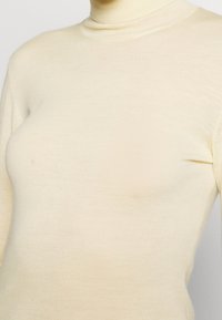 Gros plan sur une personne portant un pull à col roulé ajusté de couleur crème avec des manches longues, sur un fond uni.