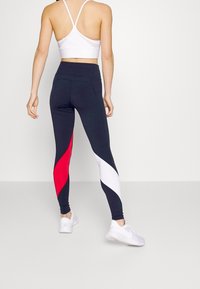 Höga leggings i marinblått med röda och vita sidopaneler; tillverkade av stretchigt tyg. Matcher med en vit sport-bh och vita sneakers.