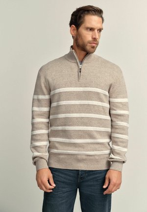 Man wearing beige quarter-zip sweater with white horizontal stripes and blue jeans, looking to the side against a plain background.