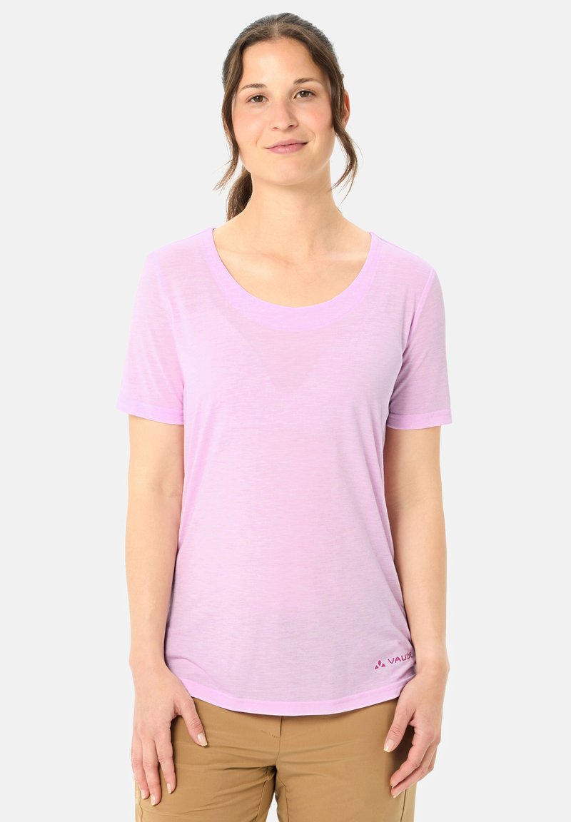 Giovane donna con capelli raccolti indossa una maglietta rosa chiaro a maniche corte e pantaloni beige, in piedi contro uno sfondo bianco uniforme.