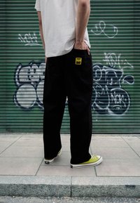 Pantalon noir à jambes larges avec une poche ornée d'une étiquette jaune, associé à des baskets basses vertes, porté avec un t-shirt blanc, devant un graffiti.